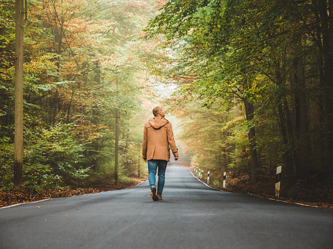Photo of single man walking or leaving through a forest landscape in a country road during autumn day. Rear view of Man walking through woods with light ahead,  after the rain stopped and the sun now shines through the forest.