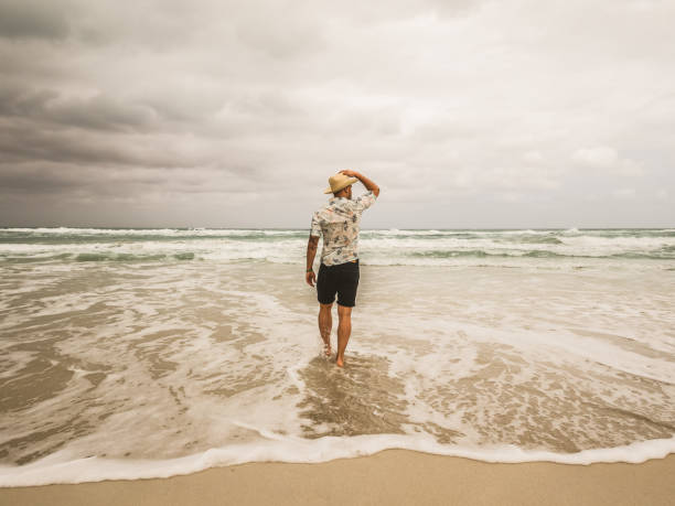 Photo of handsome man in swimming trunks standing on exotic Caribbean beach and looking out to sea, Cuba, the Caribbean, Central America. Rear view of a male enjoying the view at the beach during cloudy day.