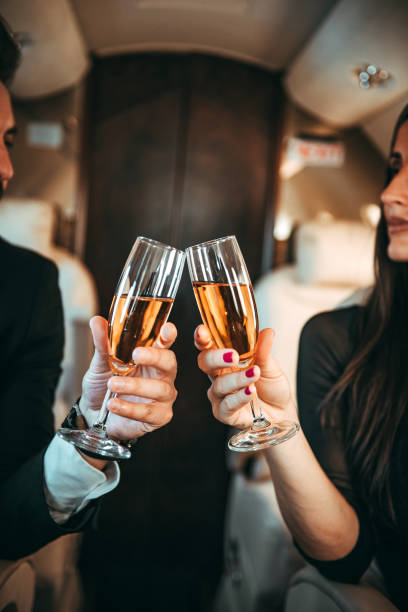Two people raising their champagne glasses in a celebratory toast aboard a private jet Young rich, well-dressed couple traveling in a private jet, drinking champagne and making a toast. Laptop and tablet are on their respective tables. private jet plane stock pictures, royalty-free photos & images