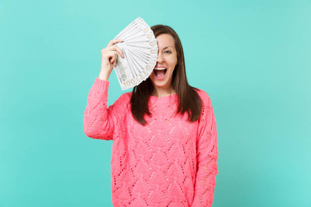 Excited young woman in pink sweater covering eye with lots bunch of dollars banknotes, cash money, keeping mouth wide open isolated on blue background. People lifestyle concept. Mock up copy space excitada a mujer suéter rosa que cubre el ojo con montón de un montón de billetes de dólares, dinero en efectivo, manteniendo boca abierto aislado sobre fondo azul. concepto de estilo de vida de las personas. se burlan de espacio de copia.