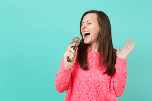 Attractive young woman in knitted pink sweater with closed eyes hold in hand, sing song in microphone isolated on blue wall background, studio portrait. People lifestyle concept. Mock up copy space mujer joven atractiva en punto jersey rosa con ojos cerrados se sostenga en la mano, cantar canto en micrófono aislado sobre fondo de pared azul, retrato de estudio. concepto de estilo de vida de las personas. se burlan de espacio de copia.