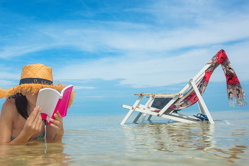 asian young woman enjoy reading a book laying on the sea water of beach, relax and happy comfort in holidays vacation sunbath on the sea beach with deck beach chair along