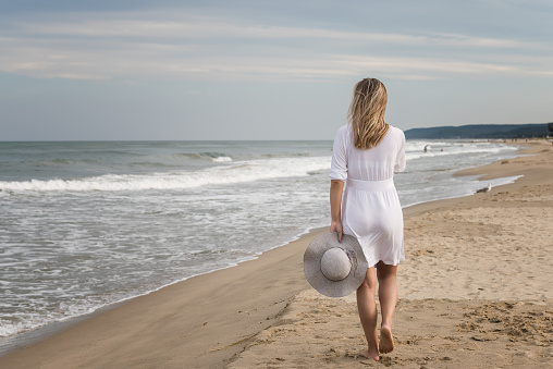 Rear view of a beautiful young woman in white dress and summer hat walking on the sandy beach