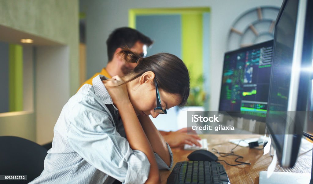 Exhausted by office work. Closeup side view of couple of mid 20's software developers resolving some issues with the code they're currently working on. The woman is in focus and she's experiencing some neck and back pain. 30-39 Years Stock Photo