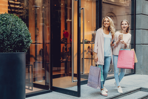 Full length portrait of charming middle-aged woman holding cellphone while enjoying shopping with daughter. They looking away and smiling