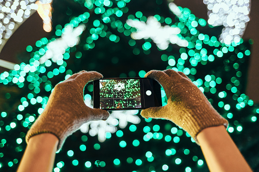 Close-up woman's hands in warm gloves taking picture of illuminated green Christmas tree using mobile phone in city at night.