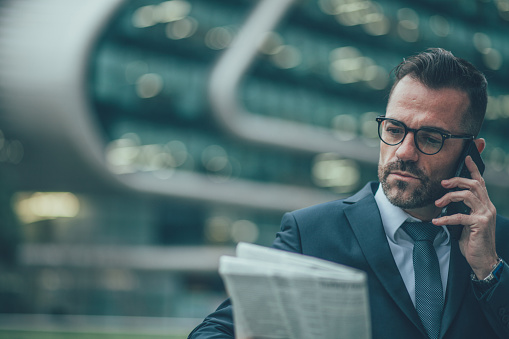 One man, handsome mature businessman, reading newspaper outdoors in city, talking on mobile.