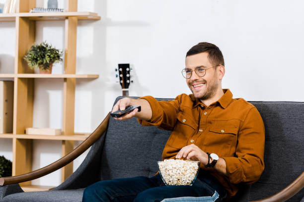 istock handsome man sitting on sofa with popcorn in bowl and holding remote control 1092700760