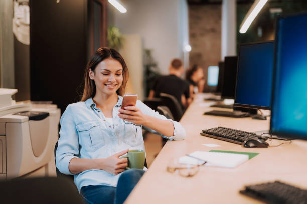 belle jeune femme avec une tasse de boisson chaude à l’aide de smartphone au travail - desk on the phone sitting table photos et images de collection