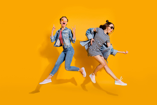Full legs body size beautiful gorgeous adorable two lady isolated on yellow background in trendy casual street style stylish denim jeans wear glasses spectacles give heavy metal gesture play guitar