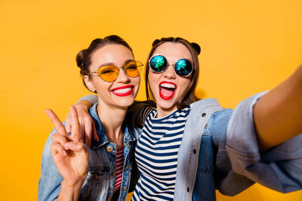 twee blij positieve grijnzende dame staan in glazen bril streetstyle cool casual stijlvol trendy denim jeans kleding geïsoleerd op gele achtergrond in nemen foto op mobiele maken hollywood smile - jong volwassen fotos stockfoto's en -beelden