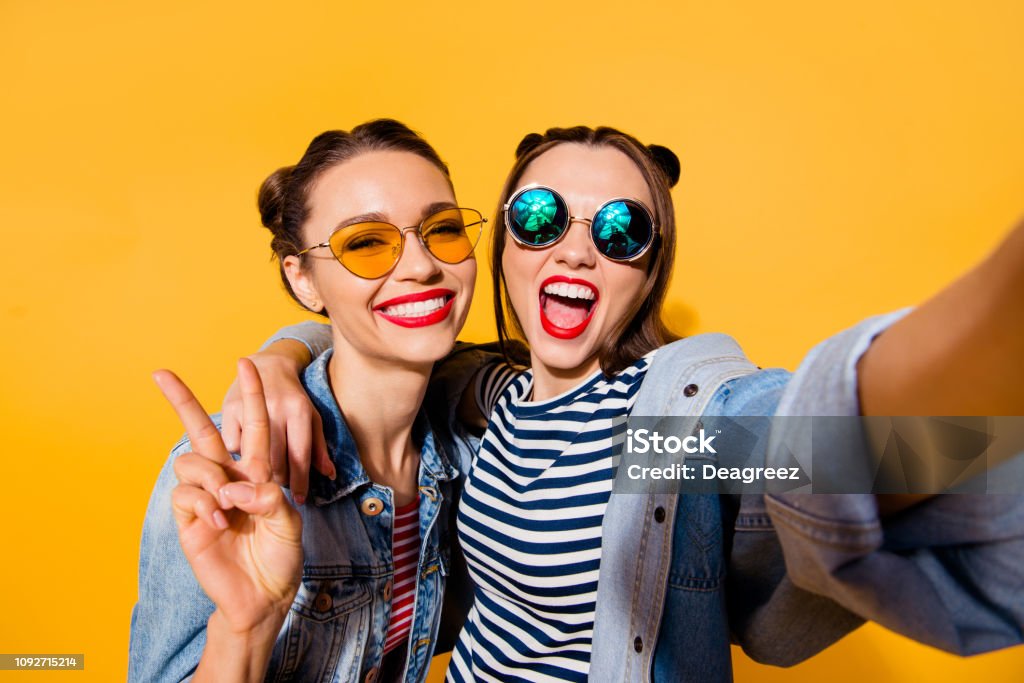Two glad positive grinning lady stand in glasses spectacles street style stylish trendy cool casual denim jeans clothes isolated on yellow background in take picture on cellular make hollywood smile Friendship Stock Photo