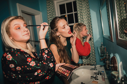 Three young women are applying makeup in the bathrooms before a night out.