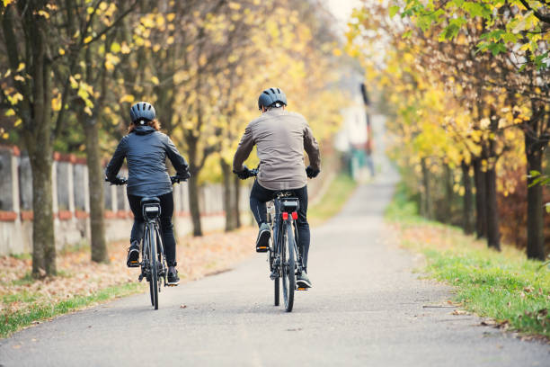 A rear view of senior couple with electrobikes cycling outdoors on a road. A rear view of active senior couple with electrobikes cycling outdoors on a road. motor living stock pictures, royalty-free photos & images