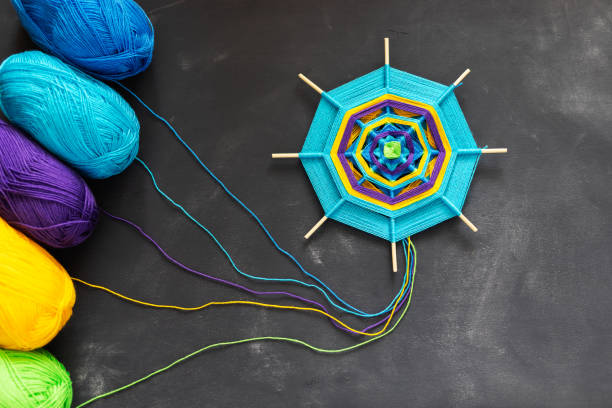 Handmade mandala of multicolor blue, purple, yellow yarn threads on dark grey background.