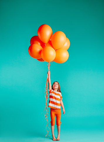 Cute girl holding orange balloons on blue background. Full length of girl child with bunch of helium balloons.