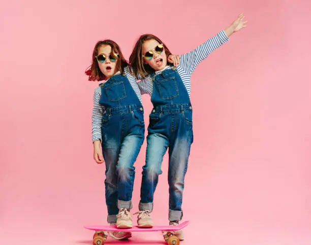 Stylish cute girls with skateboard and wearing fashion clothes on pink background. Happy children with skateboard enjoying together.
