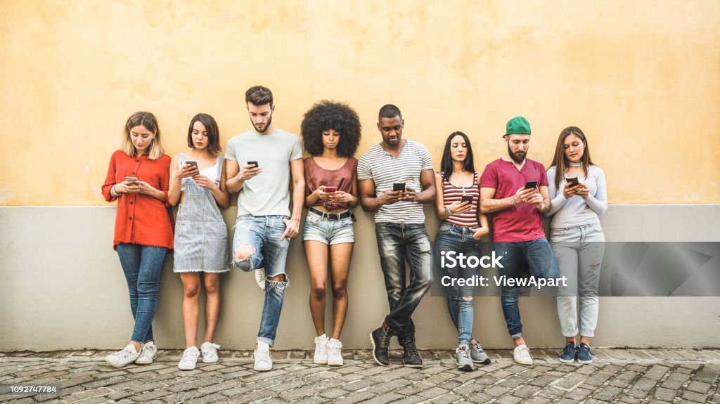 Multiracial friends using smartphone against wall at university college backyard - Young people addicted by mobile smart phone - Technology concept with always connected millennials - Filter image Generation Z Stock Photo