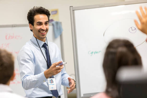 Confident male high school teacher Smiling mid adult male high school teacher points to a student with a raised hand. The student is asking or answering a question.  The teacher is holding a dry erase marker. A whiteboard is in the background. teenage high school girl raising hand during class stock pictures, royalty-free photos & images