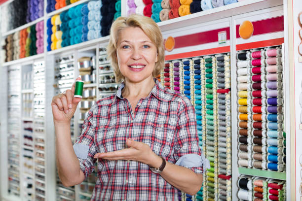 Portrait of mature cheerful female customer standing next to shelf with threads for sewing at the shop