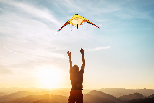 Man starting to fly bright kite in sunset sky over the mountain. Successful startup concept image.