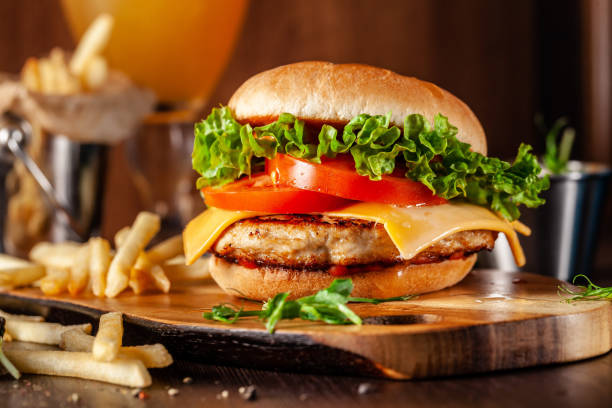 american cuisine concept. juicy burger with meat patty, tomatoes, cheddar cheese, lettuce and homemade bun. in the background are french fries and a glass of beer. close up - beer hamburger american culture beef imagens e fotografias de stock