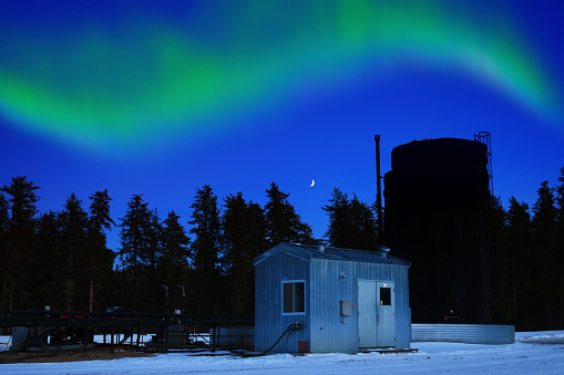 Oil lease with a bitumen tank illuminated by a moon lit sky and green northern lights