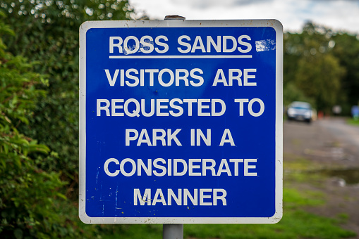 Sign: Ross Sands, Visitors are requested to park in a considerate manner, with a parked car in the blurry background - Seen in Ross Sands, Northumberland, England, UK