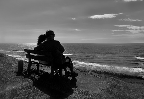 Couple sitting on bench in front of the sea, resting hugging and looking calmly forward