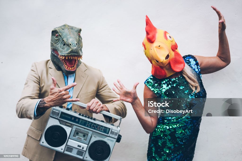 Casal sênior doido dançando para reveillon party vestindo máscara de t-rex e frango - idosos na moda se divertindo ouvindo música com boombox estéreo - conceito de tendência absurda e engraçada - focar rostos - Foto de stock de Esquisito royalty-free