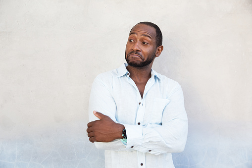 Handsome african american man showing surprise emotion. He stands in white shirt with grey blue textured wall. Casual style.