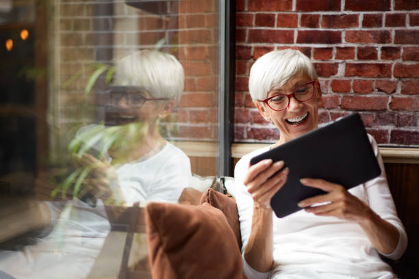 anziana donna che usa un tablet digitale e si diverte mentre è seduta comodamente accanto a una finestra di vetro - ipad senior adult facebook sofa foto e immagini stock