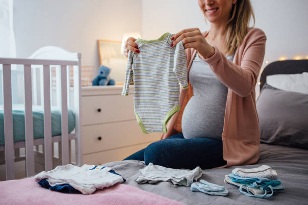 Future mom holds baby clothing Pregnant young woman holding baby clothing, sitting on bed next to the crib and smiling baby clothing stock pictures, royalty-free photos & images