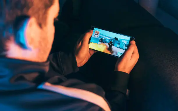 Man holds mobile in his hands and play a mobile action game. It features a crosshair and a machine gun. He sits at home at night playing the game.