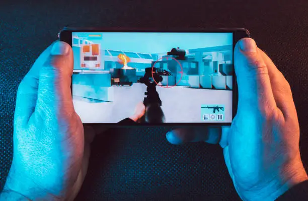 Man holds mobile in his hands and play a mobile action game. It features a crosshair and a machine gun.