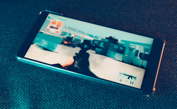Mobile phone lays on couch. It shows a sequence from an action game. It features a crosshair and a machine gun.