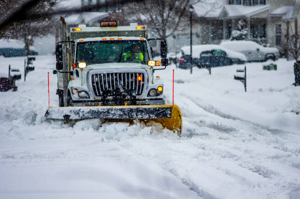 White snowplow service truck with orange lights and yellow plow blade clearing residential roads of snow while flakes are still falling Heavy equipment driver working to push snow to the side of the streets after a blizzard absence stock pictures, royalty-free photos & images