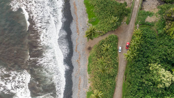 vistas aérea drone de un tramo de la carretera de hana famoso sur de hana en el lado oriental de la isla de maui, hawaii. hermosa playa de arena negra, oleaje, selva y dos coches. - hana fotografías e imágenes de stock