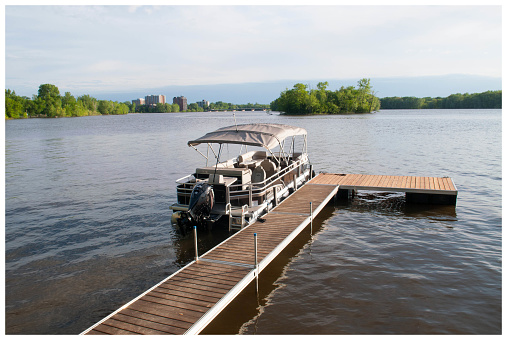 The Poonton boat was taken on the Milles-ïles river in Laval, Québec Canada.  The season is summer, the trees are green.  The pootoon boat is mooring at the cedar floating dock, in front of the dock there is a small island.  The boat is having a Bimini top.