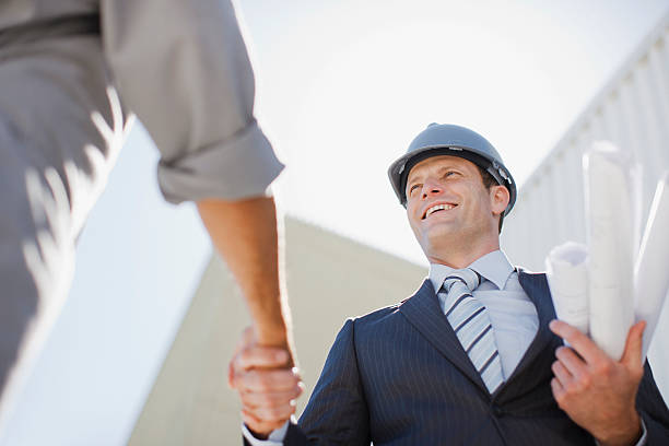 hombres de negocios estrechándose las manos con blueprints con trabajador - hardhat construction men handshake fotografías e imágenes de stock