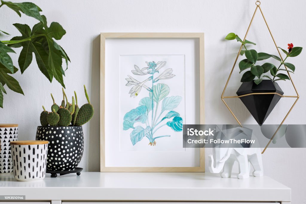 Home interior floral poster mock up with vertical wooden photo frame, a lot of plants, cacti, hanging plant in geometric pot on white wall background. Concept of white shelf. Interior floral poster mock up with vertical wooden frame. Concept with navy blue shelf Picture Frame Stock Photo