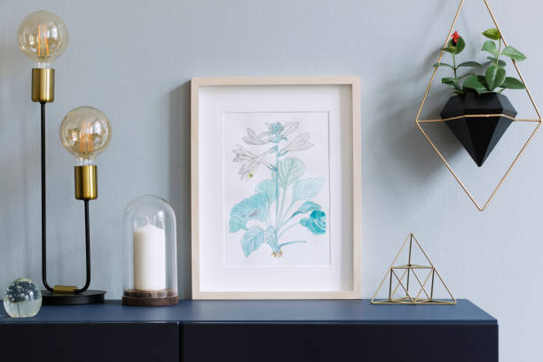 istock Interior floral poster mock up with vertical wooden frame, table lamp, accessories and hanging plants in geometric pot on the grey wall background. Concept with navy blue shelf. 1093510486