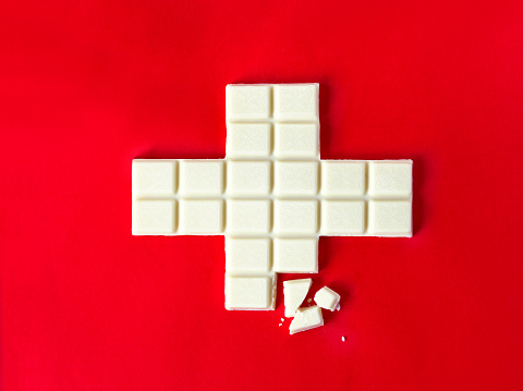 Swiss flag cross in form of white chocolate with some pieces over the red background