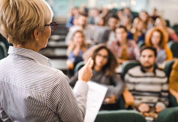 Rear view of mature teacher giving a lecture in a classroom. Back view of a senior professor talking on a class to large group of students. showing stock pictures, royalty-free photos & images