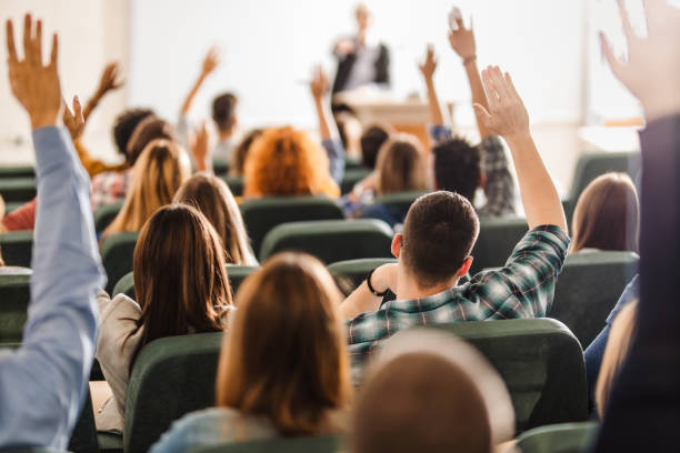 Rear view of large group of students raising arms during a class at amphitheater. Back view of college students raising their arms on a class at lecture hall. classrooms stock pictures, royalty-free photos & images