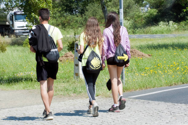 Melide, A Coruna, Spain- June 22, 2018: Rear view of three young pilgrims with small rucksackswith the symbols scallop and yellow arrow in the 'camino de Santiago' near the city of Santiago de Compostela, Galicia, Spain. The camino de Santiago is an ancient pilgrimage route which belongs to the UNESCO world heritage.