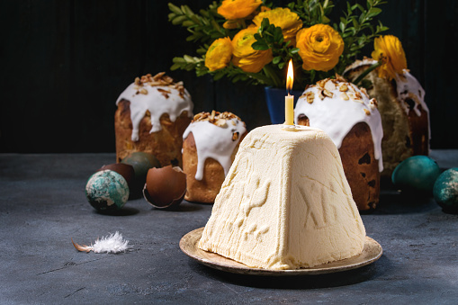 Traditional Russian Easter cottage cheese dessert, Orthodox Paskha with burning candle standing in ceramic plate on dark texture table with kulich cakes, flowers, colored eggs. Rustic style.