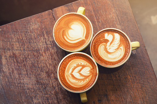 Top view of three hot cappuccino coffee in a yellow cup with various type latte art on wooden table background. (Vintage colored tone)