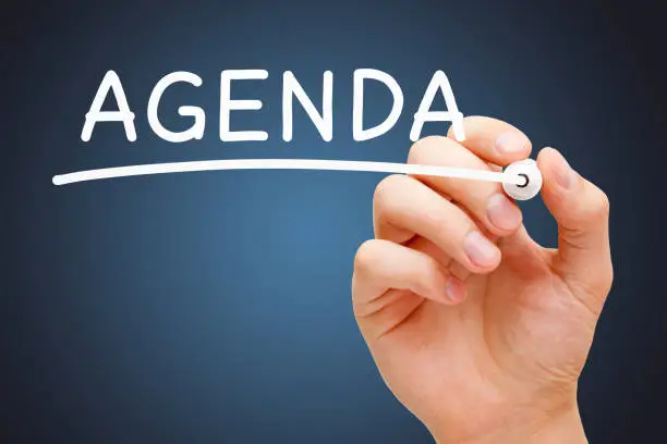 Hand writing the word Agenda with white marker on transparent wipe board over dark blue background.