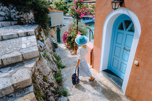 Travel tourist blonde woman with sun hat walking through narrow streets of an old greek town to the beach. Vacation summertime perfect summer day joyful holidays fun. Greece, Europe.